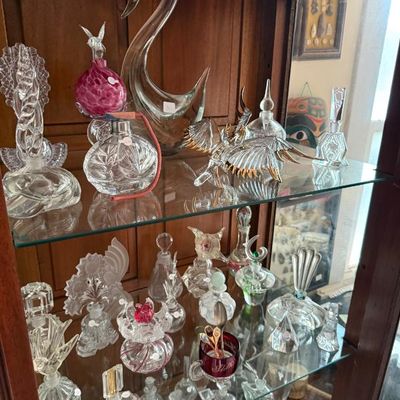 Sale Photo Thumbnail #255: Collection of vintage glass perfume bottles, vanity items, and art glass figurines, including a large swan and gold-trimmed eagle, displayed in a wooden curio cabinet.