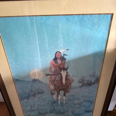 Sale Photo Thumbnail #78: Framed Western art print depicting a Native American rider on horseback holding a rifle, set against a blue sky with a low sun, presented in a dark wood frame with matting.
