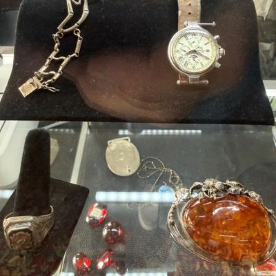 Sale Photo Thumbnail #38: Assorted fine and costume jewelry displayed in a glass case, featuring a moon phase wristwatch, a silver link bracelet, a class-style ring, and a large amber cabochon brooch.