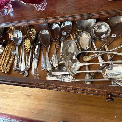Sale Photo Thumbnail #321: Large assortment of vintage silver-plated and gold-toned serving utensils, including ladles, serving spoons, and pie servers, displayed inside a wooden drawer.