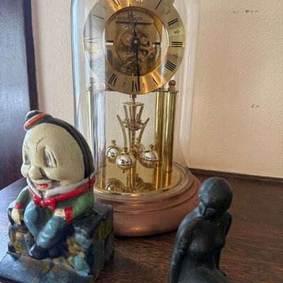Sale Photo Thumbnail #361: Vintage brass Elgin anniversary clock under a glass dome, flanked by a painted Humpty Dumpty cast iron bank and a dark metal seated mermaid figurine.