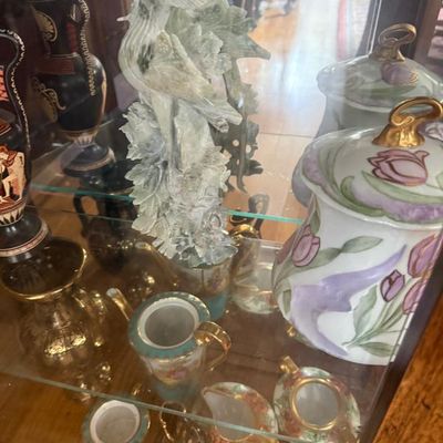 Sale Photo Thumbnail #283: A collection of vintage decorative items inside a glass display cabinet, featuring a carved jade bird sculpture, floral ceramic jars, and Greek-style black-figure urns.