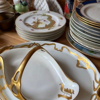 Sale Photo Thumbnail #11: Collection of vintage porcelain items, featuring a white and gold monogrammed basket dish, a stack of dragon motif plates, and decorative dishware.