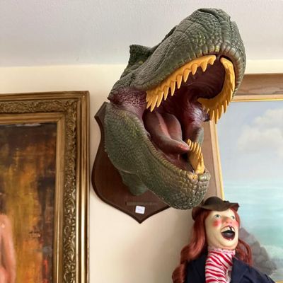 Sale Photo Thumbnail #266: Wall-mounted T-Rex dinosaur trophy head on a wooden plaque, displayed alongside a vintage doll and framed artwork.