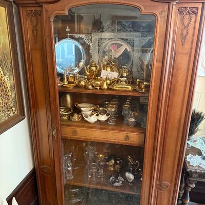 Sale Photo Thumbnail #350: Vintage French Provincial style carved wood curio cabinet with arched glass front door, displaying gold-tone tea sets, glassware, and decorative china.