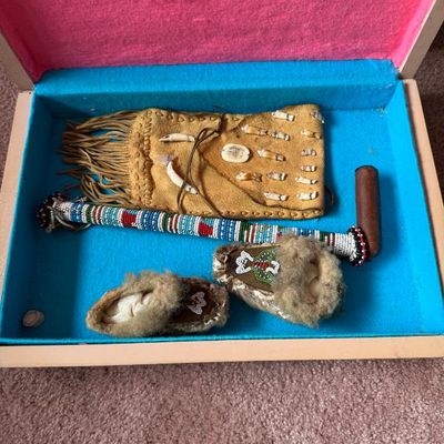 Sale Photo Thumbnail #206: Collection of Indigenous-style decorative items in a felt-lined box, including a beaded pipe, a fringe pouch adorned with shells and teeth, and a pair of small fur-lined moccasins.