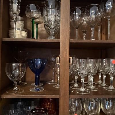 Sale Photo Thumbnail #307: Wooden display cabinet filled with an extensive collection of glassware, including cut crystal wine glasses, cobalt blue goblets, and green-stemmed drinkware.
