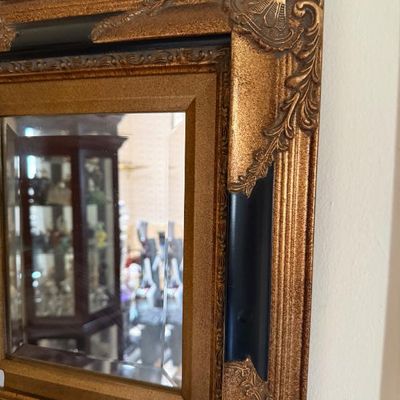 Sale Photo Thumbnail #278: Ornate wall mirror featuring a bevel-edged glass mirror set inside a composite gold-gilt and dark accent frame with shell-motif corner flourishes.
