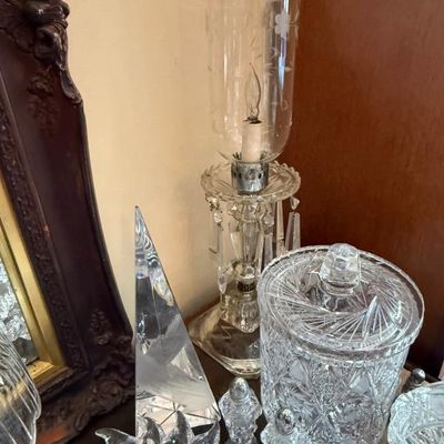 Sale Photo Thumbnail #339: Collection of vintage cut glass items, featuring an electrified hurricane table lamp with hanging crystal prisms alongside a covered jar and a frosted sailboat obelisk paperweight.