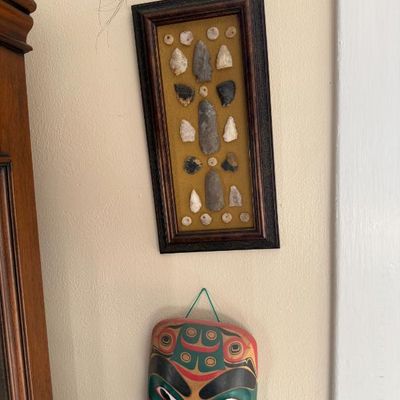 Sale Photo Thumbnail #213: Framed display of stone arrowheads and artifacts mounted on gold fabric alongside a carved Indigenous-style wooden mask on a wall.