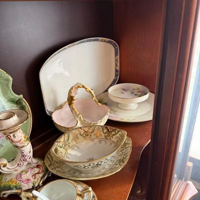 Sale Photo Thumbnail #332: Assorted vintage porcelain dishes displayed inside a wooden hutch, featuring gilded trim, hand-painted floral motifs, and decorative serving pieces.