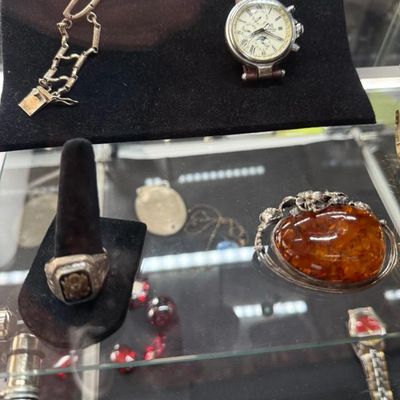 Sale Photo Thumbnail #40: Jewelry display featuring a vintage chronograph wristwatch with a leather strap, a sterling silver chain bracelet, a large amber brooch, and a military-style signet ring.