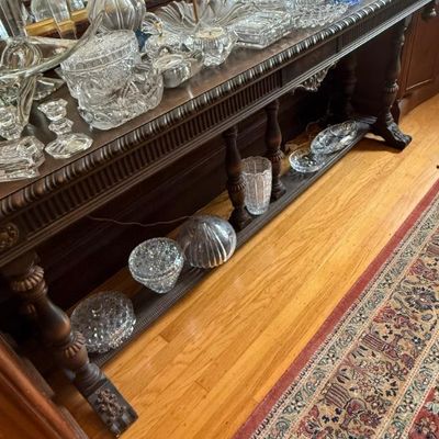 Sale Photo Thumbnail #343: A large collection of clear and cut crystal glassware, decanters, and serving pieces displayed atop and below a dark wood traditional console table.