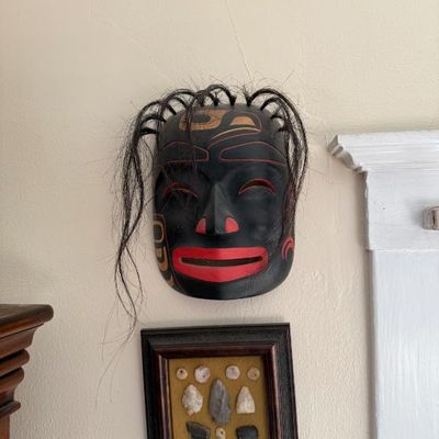 Sale Photo Thumbnail #212: Pacific Northwest Coast Indigenous-style carved wooden face mask featuring black paint with red and gold formline details, synthetic hair accents, and slit eyes, mounted on a wall above a framed arrowheads collection.