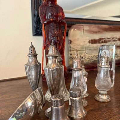 Sale Photo Thumbnail #18: Collection of vintage silver-plated and glass salt and pepper shakers displayed alongside decorative objects on a wooden table.