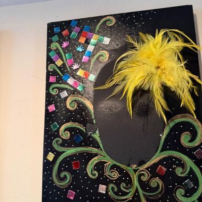 Sale Photo Thumbnail #114: Handcrafted mixed-media canvas artwork featuring a central mask-like silhouette adorned with bright yellow feathers, green swirls, and colorful mosaic tiles.