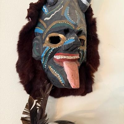 Sale Photo Thumbnail #210: Hand-carved and painted wooden folk art devil mask featuring horns, faux fur trim, and a protruding tongue, wall-mounted alongside a feather attachment.