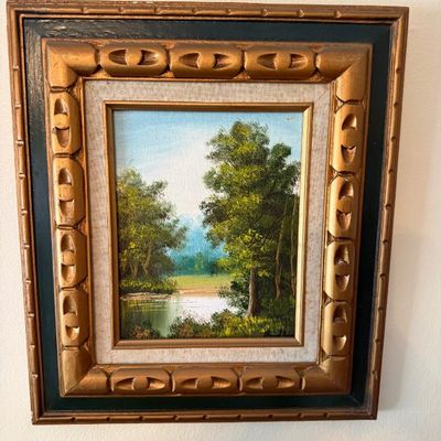 Sale Photo Thumbnail #93: Framed original oil landscape painting depicting a serene river scene surrounded by lush trees, housed in an ornate carved gilt and black frame.