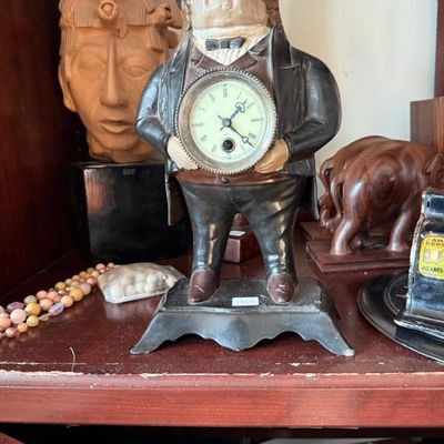 Sale Photo Thumbnail #355: Antique novelty cast iron clock depicting a gentleman holding a pocket watch face, shown alongside carved wooden decorative figures and bead necklaces.