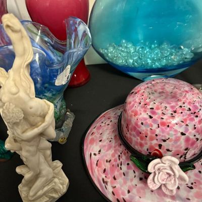 Sale Photo Thumbnail #161: Collection of decorative glass items including a pink splatter art glass hat with a flower detail, a large bright blue round vase, a red glass vase, and a classical-style figurative sculpture.