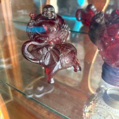 Sale Photo Thumbnail #280: Collection of carved red amber resin figurines, including an ornate trunk-up elephant and a laughing Buddha, displayed inside a glass cabinet.
