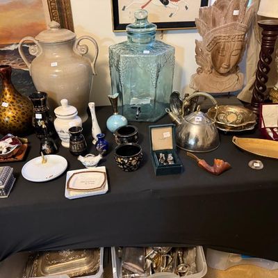 Sale Photo Thumbnail #166: Collection of eclectic home decor items displayed on a table, including a large blue glass beverage dispenser, ceramic vases, a metallic tea kettle, boxed silver novelties, and a carved wooden bust.