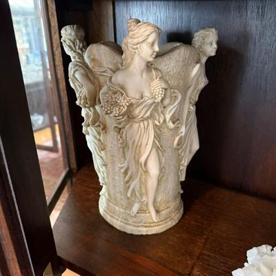 Sale Photo Thumbnail #290: Classical neoclassical-style cast resin or faux-ivory vessel featuring carved relief figures of draped maidens holding grapes, displayed inside a wooden cabinet.