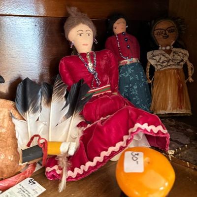 Sale Photo Thumbnail #262: Collection of vintage Native American and folk art dolls in traditional attire displayed on a shelf alongside a feather headdress accessory.