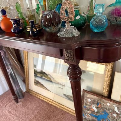 Sale Photo Thumbnail #273: Traditional dark wood console table displaying a large collection of vintage art glass and perfume bottles, with framed artwork resting against the legs below.