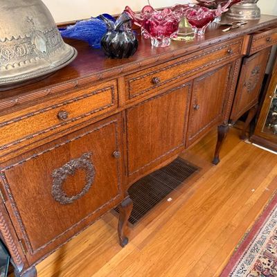 Sale Photo Thumbnail #325: Antique carved oak buffet sideboard featuring cabriole legs and ornate molded detailing, displaying a large silver cloche and decorative glass items.