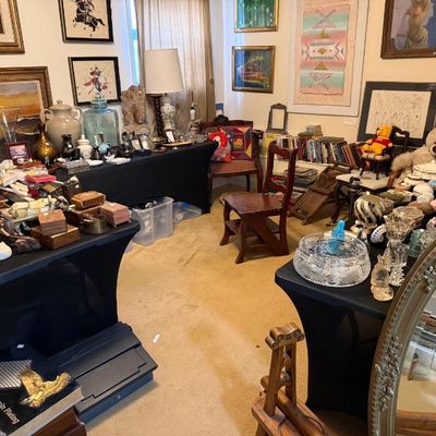 Sale Photo Thumbnail #156: A room displaying a variety of estate sale items, including framed artwork, cut glass, pottery, wooden chairs, plush toys, and collectible decor arranged on black-covered tables.