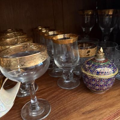 Sale Photo Thumbnail #346: Collection of vintage gold-rimmed glassware paired with a decorative Benjarong-style porcelain lidded bowl on a wooden shelf.