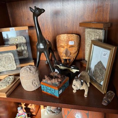 Sale Photo Thumbnail #198: Collection of global decor items on a wooden shelf, featuring a mid-century bronze-finish giraffe figurine, carved wooden mask, small bulldog statue, and framed relief plaques.