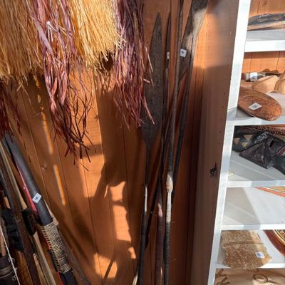 Sale Photo Thumbnail #224: Collection of tribal spears, wooden bows, and raffia grass skirts, alongside displays of carved artifacts.