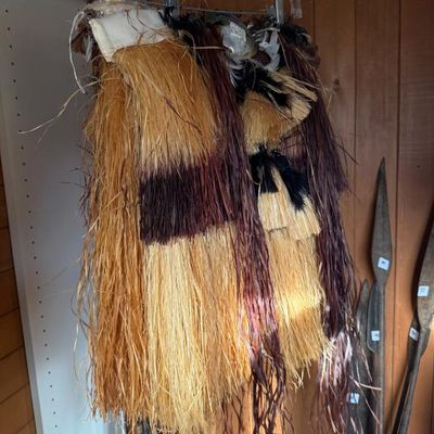 Sale Photo Thumbnail #225: Traditional Indigenous grass skirt hanging on a plastic hanger, featuring layered natural fiber fringes in natural, dark brown, and black tones.
