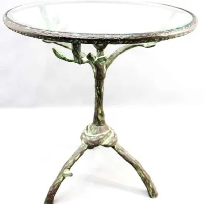 Cast metal accent table with a tree-branch form tripod base, verdigris patina, and a clear glass top.
