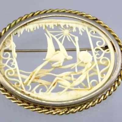 Vintage carved bone or celluoid oval brooch depicting a coastal beach scene, set in a gold-tone twisted rope and silver-tone bezel frame.