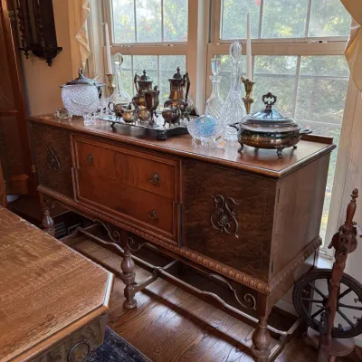 Sale Photo Thumbnail #21: Antique Jacobean-style wood sideboard buffet topped with silver-plate tea service, crystal decanters, and serving pieces, flanked by a wall clock and spinning wheel.