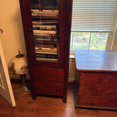 Sale Photo Thumbnail #125: Tall dark wood display cabinet featuring a glass pane door, internal shelving filled with books, and two lower drawers.