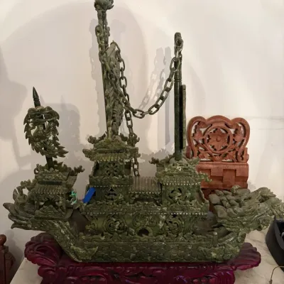 Sale Photo Thumbnail #43: Ornately carved green jade or serpentine dragon boat sculpture resting on a fitted reddish wooden stand, featuring intricate pierced lattice work and linked chain details.