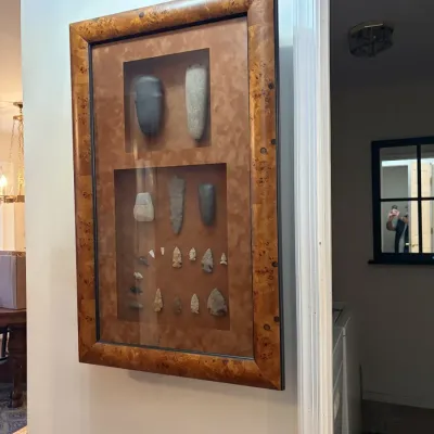 Sale Photo Thumbnail #102: Framed wall-mounted display shadow box containing a collection of Native American stone artifacts, including celts, axes, and arrowheads, set in a burl wood frame.