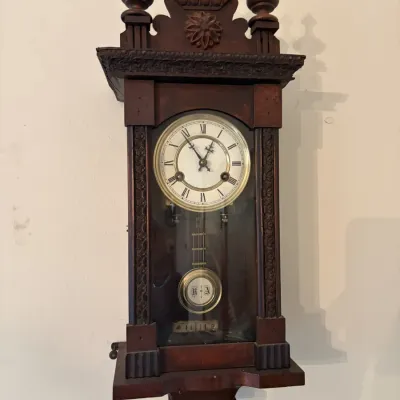 Sale Photo Thumbnail #23: Antique wooden regulator-style wall clock featuring turned finials, carved accents, and a brass pendulum behind a glass door.