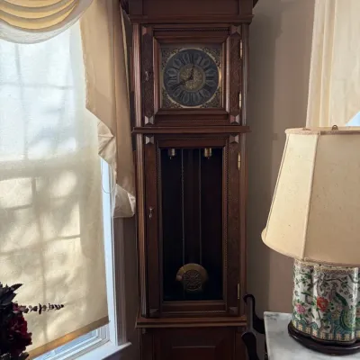 Sale Photo Thumbnail #9: Traditional wooden grandfather clock featuring a bonnet top, ornate dial, and visible pendulum through a glass door.