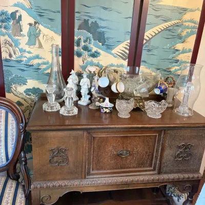 Sale Photo Thumbnail #22: Vintage Jacobean revival style wooden sideboard displaying clear glass decanters, punch bowl set, pressed glass items, and ceramic figurines, positioned in front of a painted folding screen.