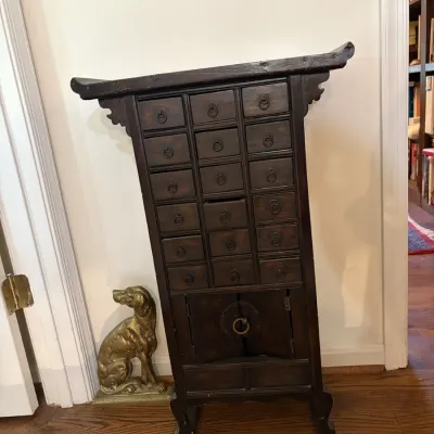 Sale Photo Thumbnail #96: Asian-style dark wood apothecary cabinet featuring multiple small drawers, a bottom double-door compartment, and pagod-style top trim, pictured beside a brass dog doorstop.