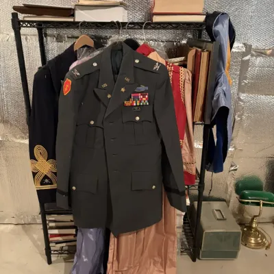 Sale Photo Thumbnail #98: Vintage US Army Class A dress green uniform jacket displaying rank insignia, ribbon bars, and a Combat Infantryman Badge, hung on a wire clothing rack alongside other garments.