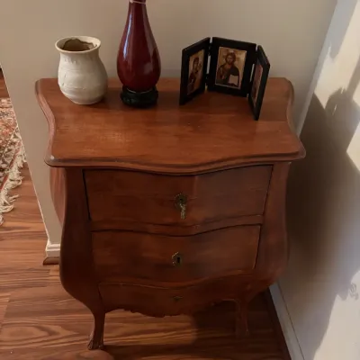 Sale Photo Thumbnail #89: French Provincial style wood bombshell nightstand with three drawers, paired with decorative items including a red glass vase, ceramic jar, and a small triptych icon frame.