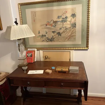 Sale Photo Thumbnail #130: Traditional wooden drop-leaf desk featuring ring pull drawers, topped with various small trinket boxes, books, and a cut crystal table lamp with a pleated shade.