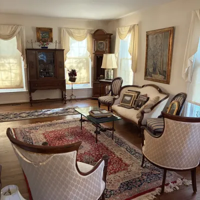 Sale Photo Thumbnail #28: Traditional living room set including a cream-upholstered camelback sofa, patterned accent armchairs, a glass-top coffee table, and a large red Oriental area rug.