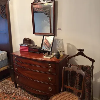 Sale Photo Thumbnail #80: Traditional mahogany bow-front dresser paired with a broken-arch pediment wall mirror, a vintage valet stand, and a cane-seat side chair.